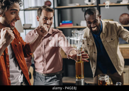 excited multiethnic men building tower from bottles and glasses while having fun together at party Stock Photo