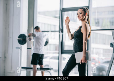 female personal trainer with digital tablet waving hand and athletic man exercising with barbell behind at gym Stock Photo
