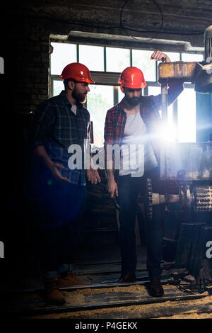 two bearded workers in protective helmets using machine tool at sawmill Stock Photo