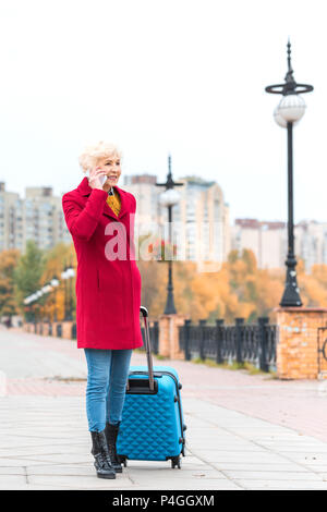 senior woman in red coat walking with suitcase and talking on smartphone Stock Photo