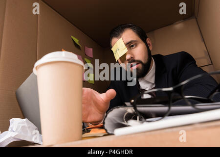 exhausted businessman with sticky note on forehead taking disposable cup of coffee in box Stock Photo