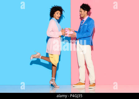Young african amercian man gives girl gift box on pink and blue background on pink and blue background Stock Photo