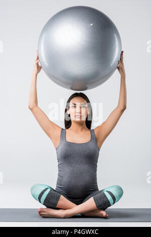 attractive pregnant asian woman sitting in lotus pose and holding fitness ball, isolated on grey Stock Photo