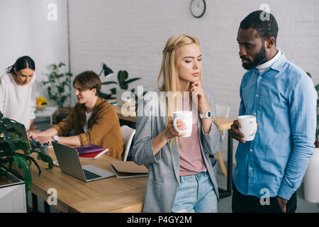 multicultural business coworkers with coffee cups having discussion and two coworkers working on laptop behind Stock Photo