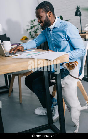 side view of smiling african american businessman working on laptop and dog standing near Stock Photo