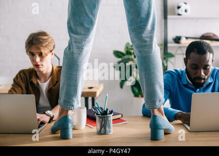 cropped image of female standing on table and two scared multiethnic businessmen working on laptops Stock Photo
