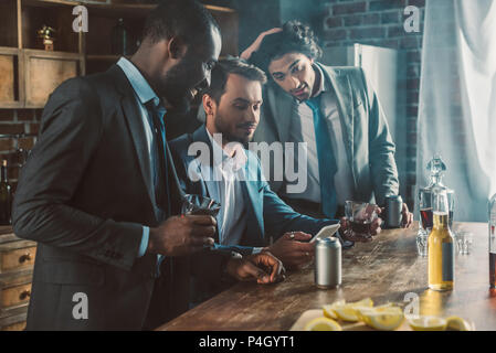 three young men in suits looking at smartphone while drinking whisky together Stock Photo