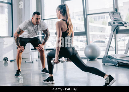 male personal trainer looking at sportswoman doing sit ups with dumbbells at gym Stock Photo