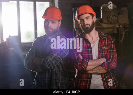 two bearded carpenters with crossed hands in protective helmets standing at sawmill Stock Photo