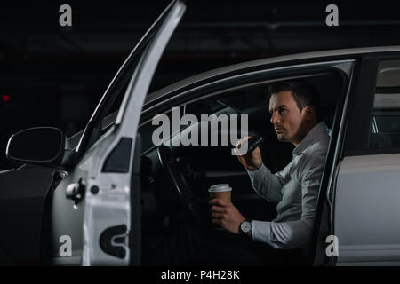 undercover male agent with cup of coffee doing surveillance by binoculars from car Stock Photo