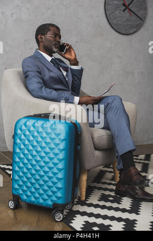 Businessman talking by smartphone and sitting in armchair, travel bag standing near Stock Photo