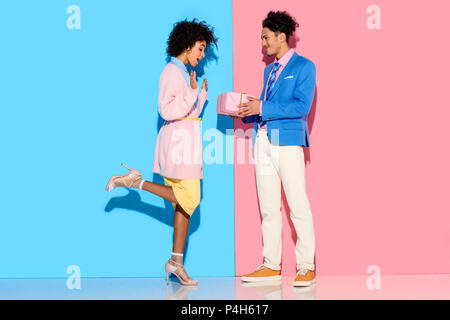 Young african amercian man gives girl gift box on pink and blue background Stock Photo