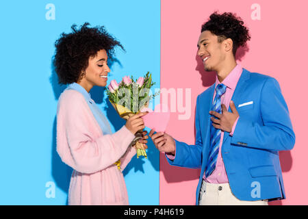 Young african amercian man gives girl bouquet of flowers and heart card on pink and blue background Stock Photo