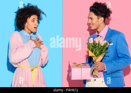 Young african amercian man gives surprised girl bouquet of flowers and gift box on pink and blue background Stock Photo