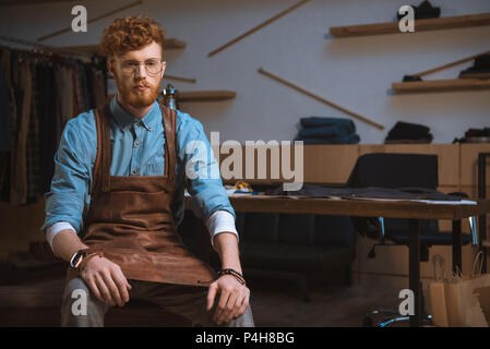 serious male fashion designer in apron and eyeglasses sitting at workshop Stock Photo