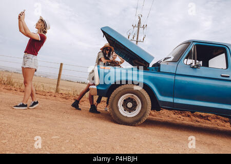 Female friends examining broken down car on country road. Woman looking under the hood of broken down car with friends searching for mobile network to Stock Photo