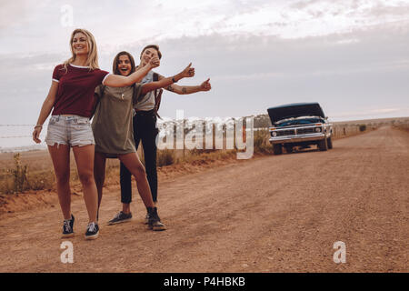 Three young women hitchhiking near their broken car on country road. Smiling female friends gesturing for a lift on rural road with broken down car in Stock Photo