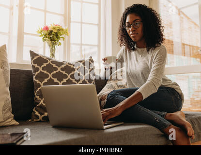 Businesswoman working on laptop computer sitting at home holding a coffee cup in hand. Woman sitting on sofa at home and managing her business. Stock Photo