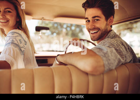 Handsome young man driving the car with his girlfriend sitting by, both looking back and smiling. Couple enjoying on a road trip. Stock Photo