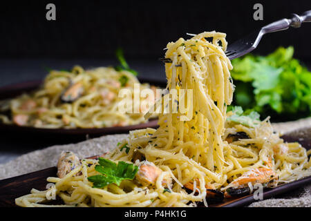 A man takes a fork with a fork from a plate on a wooden table. Pasta with seafood, prawns and mussels in creamy sauce. Photo on a dark background Stock Photo