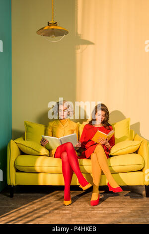 women in retro clothing reading books sitting on yellow sofa at bright apartment, doll house concept Stock Photo