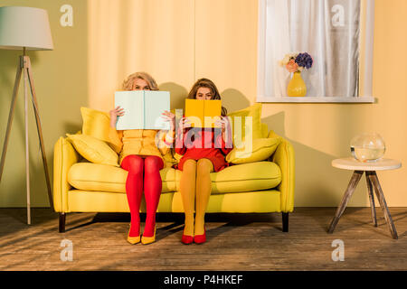 obscured view of women in retro clothing with books sitting on yellow sofa at bright apartment, doll house concept Stock Photo