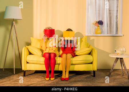 obscured view of women in retro clothing with books sitting on yellow sofa at bright apartment, doll house concept Stock Photo