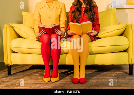 cropped shot of women in retro clothing with books sitting on yellow sofa at bright apartment, doll house concept Stock Photo