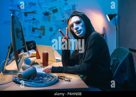 female hacker in mask showing v sign at workplace Stock Photo