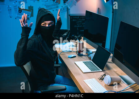 busted hacker in mask with raised hands in front of workplace Stock Photo
