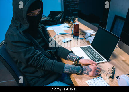busted hacker in mask with handcuffs in front of workplace Stock Photo