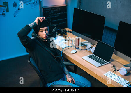 young female hacker at in front of desk taking off mask Stock Photo