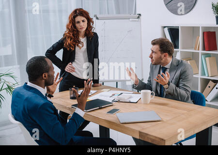 Financial adviser standing near table with laptop and papers while two businessmen arguing with her Stock Photo
