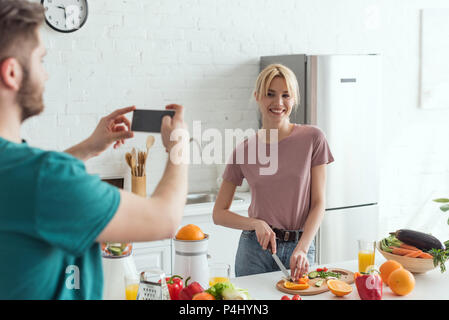 partial view of man taking picture of smiling girlfriend cooking in kitchen at home, vegan lifestyle concept Stock Photo