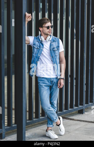 handsome young man in denim vest leaning on fence on city street Stock Photo