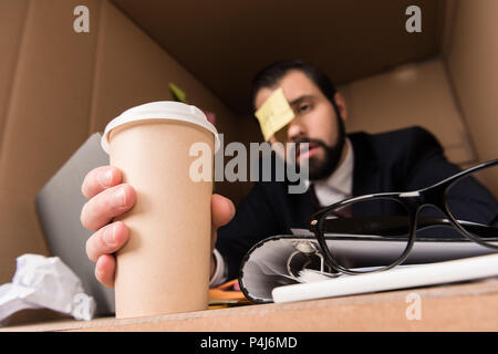 exhausted businessman with sticky note on forehead taking disposable cup of coffee in box Stock Photo