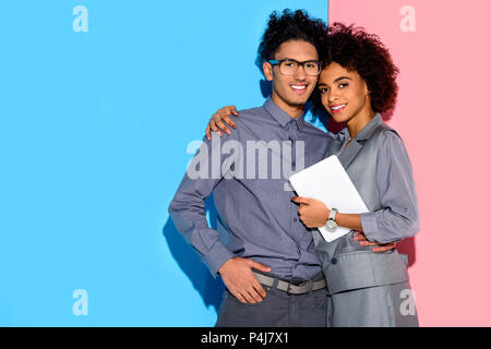 Young african amercian businesswoman holding tablet and hugging stylish businessman on pink and blue background Stock Photo