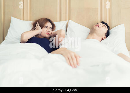 Annoyed Asian wife blocking her ears from noise of husband snoring in bedroom at home. Young couple have problem with man's snoring. Stock Photo