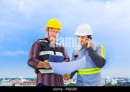 Asian construction engineer talking on the phone while working with his foreman looking at the blueprint at construction site, good for teamwork and e Stock Photo