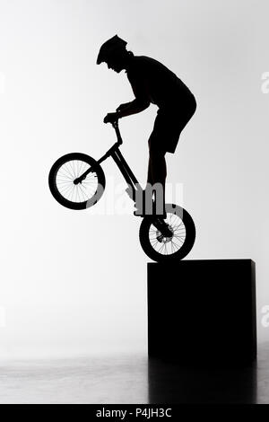 silhouette of trial biker performing stunt while balancing on cube on white Stock Photo