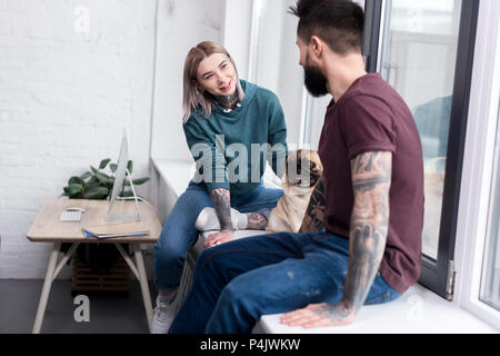 happy tattooed couple sitting on windowsill with pug at home Stock Photo