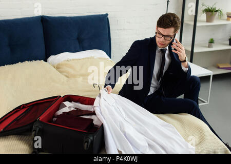 handsome businessman talking on smartphone while packing suitcase in bedroom Stock Photo