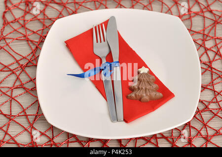 A simple place setting for Christmas consisting of a plate, fork, knife, red napkin and a chocolate shape christmas tree on a red place mat Stock Photo