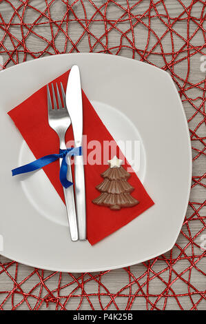 A simple place setting for Christmas consisting of a plate, fork, knife, red napkin and a chocolate shape christmas tree on a red place mat Stock Photo