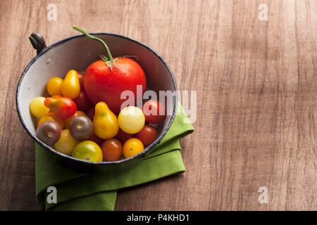 A variety of tomatoes in an antique enamel bowl on wood background with a green napkin Stock Photo