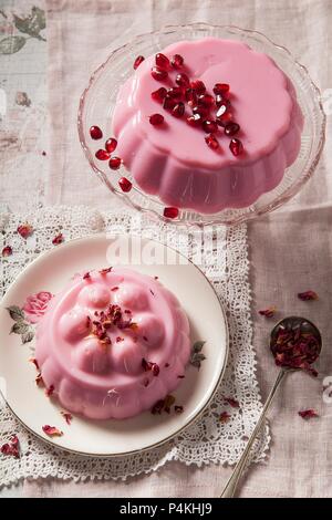 Two pastel pink milk jellies on vintage glass cake stand and plate one covered with pomegrante seeds the other with dried rose petals shot overhead on vintage lace napkin Stock Photo