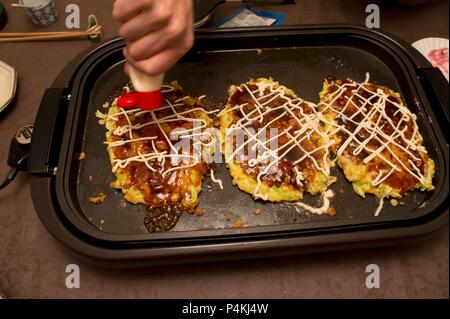 Okonomiyaki (Japanese pancakes) on a hot tray being decorated with mayonnaise Stock Photo