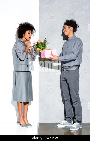 Young african amercian man gives girl a bouquet of flowers and gift on grey and white background Stock Photo