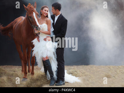 Young newlywed couple, beautiful beauty bride in fashion white bridal wedding costume and handsome groom standing by muscular male horse on black drea Stock Photo