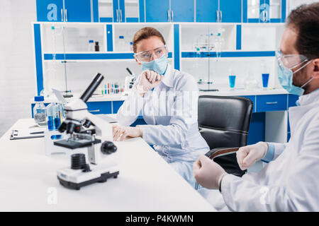 scientific researchers in medical masks and goggles having discussion at workplace with microscope in lab Stock Photo